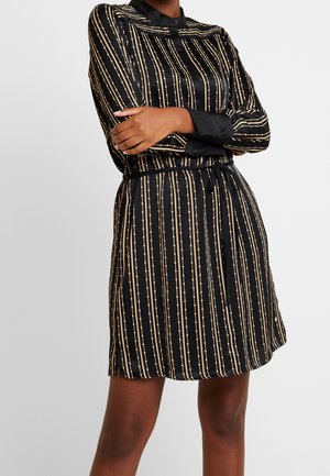 Kvinde iført en sort satin kjole med lodrette beige perle-lignende striber, lange ærmer, taljebånd og stående med korslagte arme.