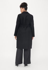Cappotto di lana nera con vita regolabile, reverse e maniche lunghe. Abbinato a pantaloni grigi a gamba larga e tacchi neri.