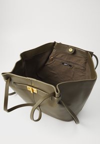 Oliivinvihreä kangaskassi teksturoidulla ulkopinnalla, jossa on kullanvärinen solki ja ruskea kangasvuori sisäisellä vetoketjutaskulla.