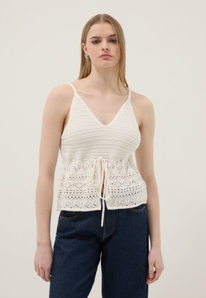 Jonge vrouw met lang blond haar, gekleed in een wit gehaakt mouwloos topje met strikdetail en donkerblauwe spijkerbroek, tegen een effen achtergrond.