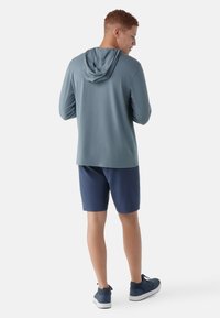 Junger Mann, der von hinten zu sehen ist, trägt ein türkisfarbenes langärmliges Kapuzenoberteil, marineblaue Shorts und dunkelblaue Sneaker vor einem weißen Hintergrund.