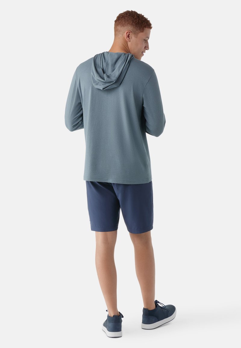 Młody mężczyzna z tyłu, ubrany w turkusową długą koszulę z kapturem, granatowe szorty i ciemnoniebieskie trampki na białym tle.