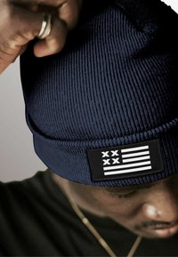 Navy blue ribbed beanie with a black patch featuring a white flag design. Textured fabric and snug fit. Close-up view with hands adjusting.