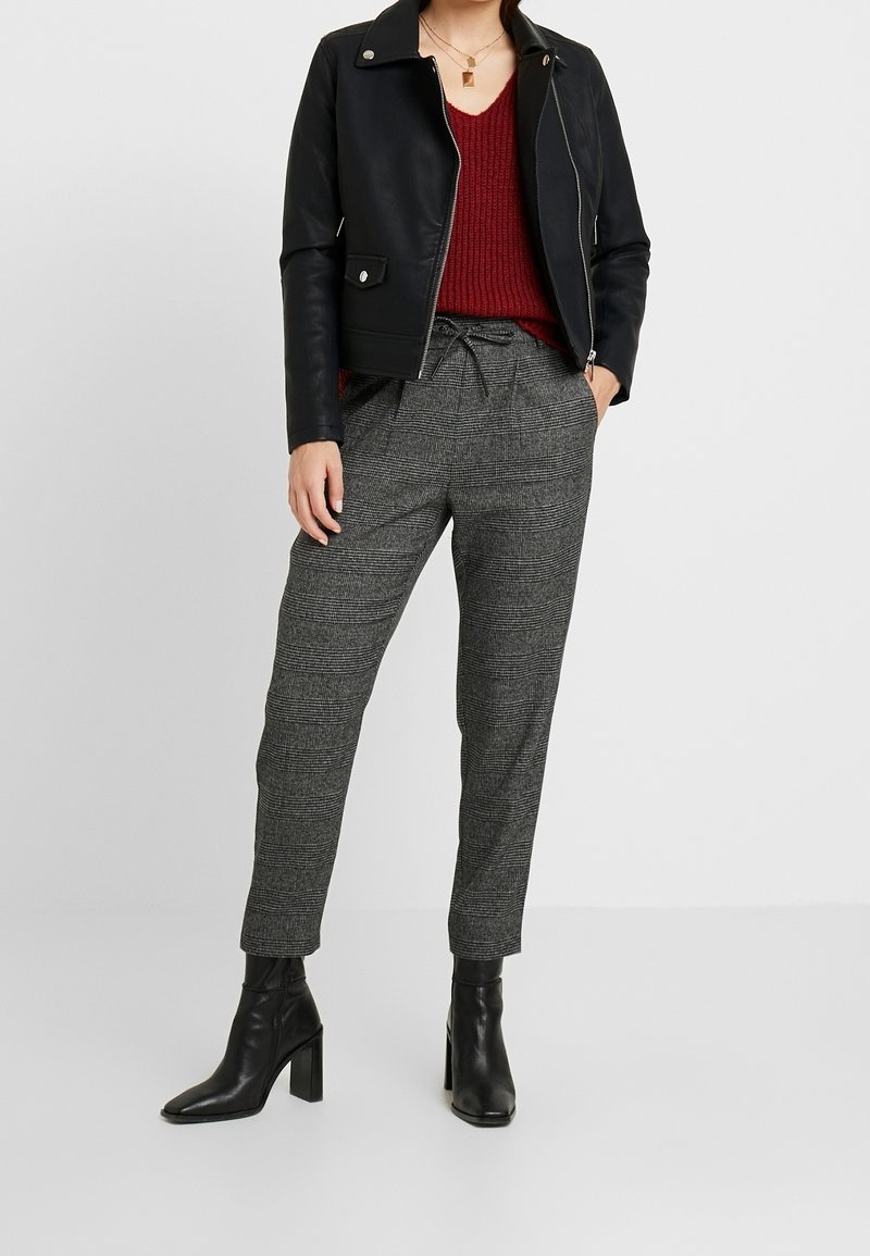 Femme portant une veste en cuir noire, un pull en maille rouge, un pantalon gris à carreaux avec cordon, des bottines noires à talons et des colliers dorés superposés.