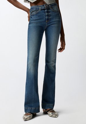 Γυναίκα με μπλε καμπάνα τζιν, φλατ παπούτσια μυτερά με φιδίσιο σχέδιο και μια μικρή μαύρη ζώνη σε λευκό φόντο.