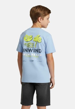 Jongen draagt een lichtblauw T-shirt met een geel ontwerp van tropische bladeren en een palmboom, en donkergrijze korte broek, staand zijwaarts naar rechts gericht.