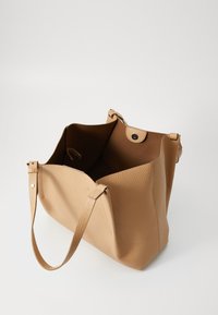 Tan leren tote bag met een geprofileerde textuur, met twee korte handgrepen en een drukknoopsluiting. De binnenkant is bruin met een ruime indeling.