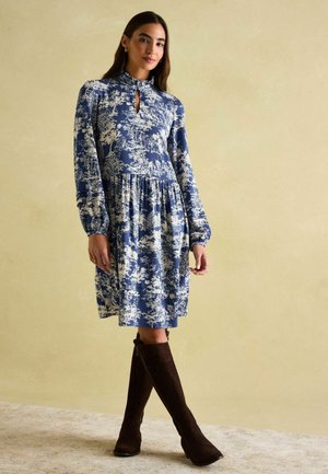 Ein blaues Blumen-Kleid mit langen Ärmeln, einem hohen Kragen und einem Schlüssel-ausschnitt, ergänzt durch braune kniehohe Stiefel. Weicher Stoff und plissierter Rock.