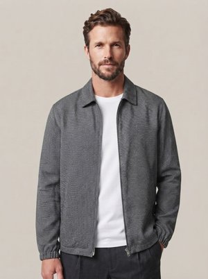 Mann mit kurzem braunem Haar und Bart, der eine graue, strukturierte Jacke mit Reißverschluss über einem weißen Hemd vor einem schlichten beigen Hintergrund trägt.