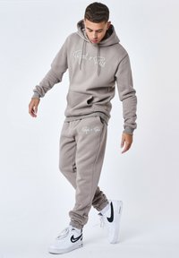 Grijze hoodie en joggingset gemaakt van zachte stof, met een kangoeroezak, een capuchon met trekkoord en witte logo-accenten.