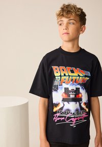 Tricou negru din bumbac, cu un grafic colorat reprezentând o mașină cu flăcări, textul "Back to the Future" și "Powered by Flux Capacitor".