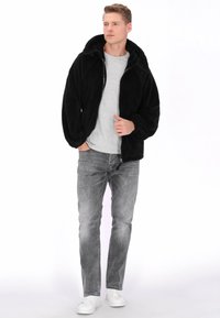 Czarna kurtka polarowa z kapturem na pełnym zapięciu, noszona na jasnoszarej koszulce. Połączona z jasnoszarymi jeansami i białymi trampkami.