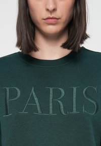 Donna con capelli scuri lunghi fino alle spalle, che indossa una maglietta verde scuro con la scritta ricamata in rilievo "PARIS" sul davanti.