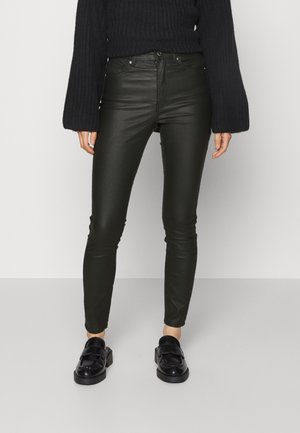 Femme portant un pantalon skinny en cuir noir, des mocassins noirs épais et un pull côtelé noir oversize, debout sur un sol gris uni.