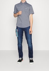 Szara, krótka koszula na guziki z kołnierzykiem i logo. Noszona z niebieskimi dżinsami i czarnymi sneakersami. Mała niebieska torebka na ramię.