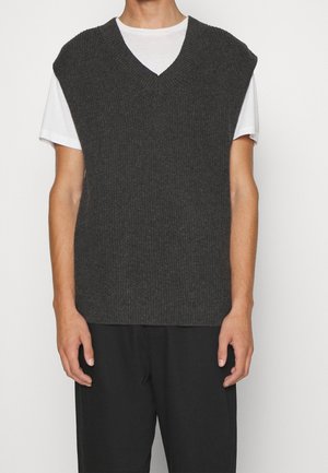 Person trägt eine dunkelgraue, gestrickte ärmellose Strickweste über einem weißen T-Shirt mit kurzen Ärmeln und schwarzen Hosen, steht vor einem schlichten Hintergrund.