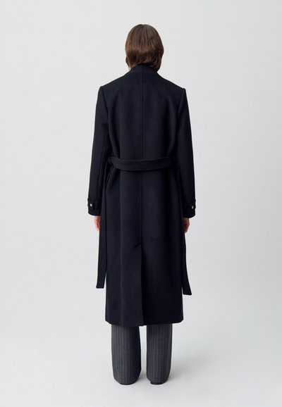 Trench coat nero in lana con cintura, forma strutturata, maniche lunghe e polsini bottonati. Abbinato a pantaloni grigi a righe.