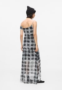 Kvinde iført en sort beanie og en sort-hvid plaid maxi-kjole med én skulder, stående sidelæns mod en hvid baggrund.
