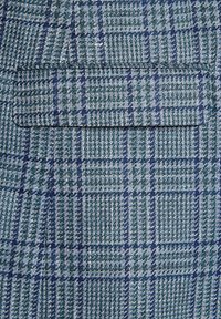 Tkanina w zielono-niebieską kratę z teksturowanym splotem; zawiera jedną kieszeń z klapką, co nadaje jej zorganizowany wygląd.