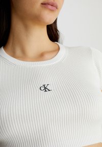 Top scurt tricotat alb cu textura ribbed, guler rotund și logo-ul brodat "cK" în negru pe partea stângă. Textură netedă și design ajustat.