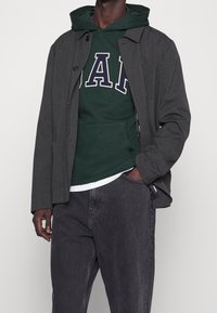 Donkergrijze buitenjas met knoopsluiting en zijzakken over een groene hoodie met witte en blauwe letters, gecombineerd met donkere spijkerbroek.