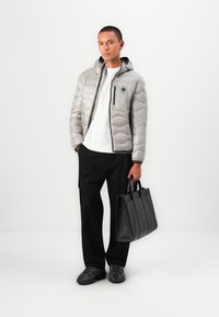 Grijze, gewatteerde pufferjack met een capuchon en rits, gedragen over een wit T-shirt. Zwarte broeken en een zwarte gestructureerde tote bag. Sneakers met textuur.