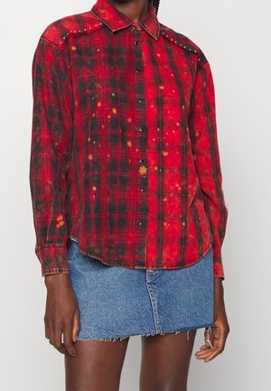 Czerwona i czarna koszula w kratę zapinana na guziki, z pomarańczowymi plamami typu tie-dye i srebrnymi ćwiekami na ramionach, noszona z niebieską spódnicą dżinsową.