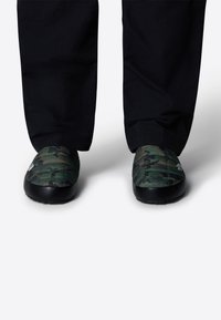 Scarpe slip-on verde mimetico con texture trapuntata e suole in gomma nera, abbinate a pantaloni neri, mostrando solo le calzature.