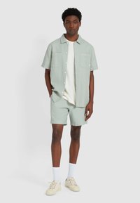 Lichtgroene getextureerde kortekostschouderhemd met twee borstzakken, gecombineerd met bijpassende shorts. Witte t-shirt eronder, beige sneakers en witte sokken.