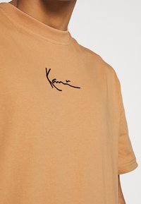 Camiseta de algodón color beige con mangas cortas, que presenta un logotipo negro en cursiva bordado en el pecho. Cuello de barco, textura suave.