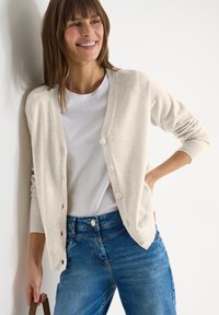 Femme souriante, portant un cardigan beige clair boutonné sur un t-shirt blanc et un jean bleu, debout contre un mur blanc, tenant une anse de sac.