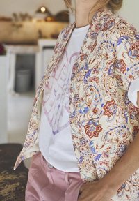 Camicia a fantasia floreale color crema, con disegni blu e rossi, indossata sopra una maglietta grafica bianca. Pantaloni corti rosa chiaro completano l'outfit.