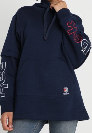 Osoba ubrana w granatową bluzę z kapturem Reebok z logo na kieszeni oraz czerwonym, białym i niebieskim napisem na obu rękawach, w połączeniu z niebieskimi dżinsami.