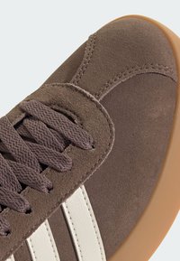 Smeđe suede tenisice s teksturiranom površinom, ljubičastim vezicama, bijelim dizajnom s tri pruge i gumenenom đonom. Krupni kadar naglašava detalje.