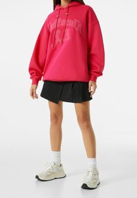 Růžová přehnaně velká mikina s šedým grafickým textem, spárovaná s černou sukní s jemným proužkem. Bílé ponožky a béžové tenisky dotvářejí celý outfit.