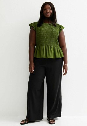 Kobieta stojąca, ubrana w zielony górny top z marszczonymi rękawami, czarne szerokie spodnie i czarne sandały na tle jednolitego białego tła.