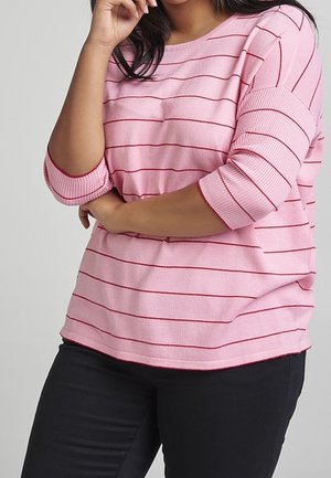 Vrouw draagt een roze shirt met lange mouwen met dunne rode horizontale strepen en een zwarte broek, staat met één hand aan haar kin.
