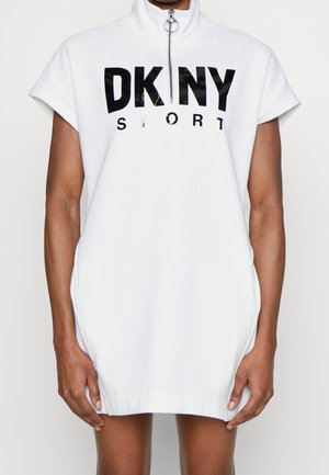 Femme portant une robe sportive blanche sans manches avec une fermeture éclair à moitié et "DKNY SPORT" imprimé en noir sur la poitrine.