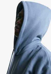 Kapucnis pulóver puha kék anyagból, húzózsinórral, laza szabású és texturált felülettel; közeli profil nézet.