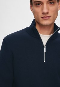 Marineblå halv-zip tröja gjord av strukturerad stickad tyg, med ribbad krage och silverfärgad dragkedjedetalj. Inga tydliga mönster eller accenter.