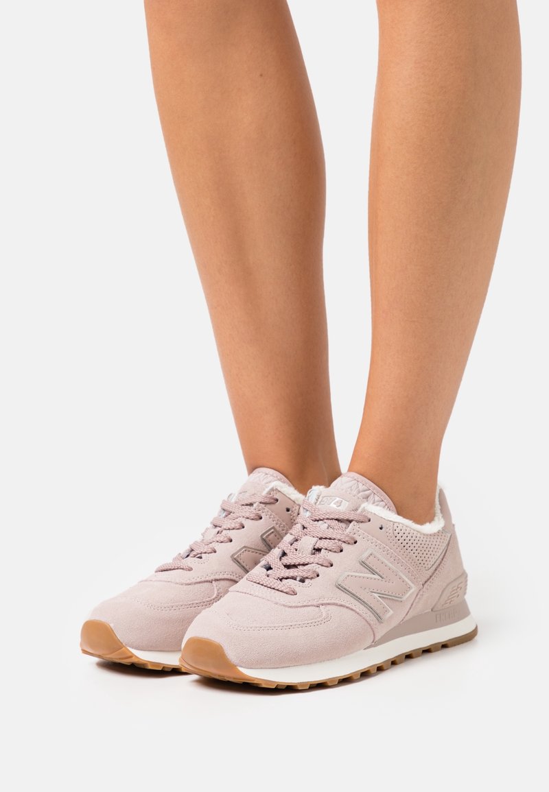 Baskets en daim rose clair avec un col en peluche blanc, des lacets texturés et une semelle en caoutchouc marron. Elles présentent un logo "N" argenté sur le côté.