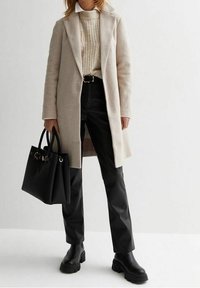 Beige villakangas takki, jossa on kaulukset, ribatusta kermanvärisestä neuleesta, mustat tekonahkahousut, mustat nilkkasaappaat ja jämäkkä musta käsilaukku.