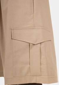 Pantaloni cargo beige con tasca laterale dotata di una patta a punta, realizzati in tessuto leggero con una superficie liscia e dettagli di cucitura ordinati.