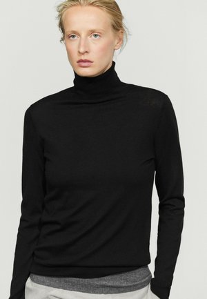 Donna dalla pelle chiara e capelli biondi raccolti, indossa un maglione nero a collo alto sopra una camicia grigia su uno sfondo chiaro uniforme.