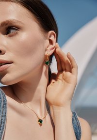 Orecchini a forma di cuore in tono oro e collana coordinata con gemme verdi, indossati da un modello con uno sfondo naturale. Texture lisce visibili.