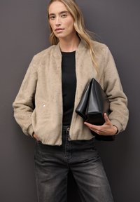 Faux fur bomberjacka i ljus beige med ribbade ärmslut och nederkant. Svart topp under, håller en vikt svart läderclutch. Mörka jeans.