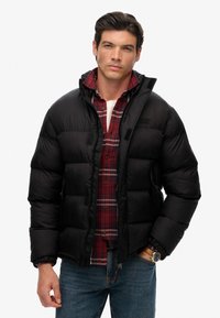 Chaqueta de plumón negra con bolsillos con cremallera, con un acabado brillante. Usada sobre una camisa de cuadros roja y azul con camiseta blanca por debajo, jeans azules.