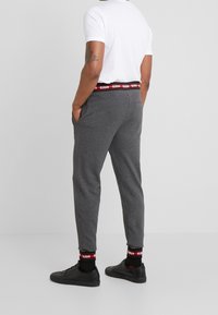 Graue Sweatpants mit einem tapered Design, ausgestattet mit seitlichen Taschen und einem schwarzen Bund mit rotem Text. Kombiniert mit schwarzen Schuhen und Socken.
