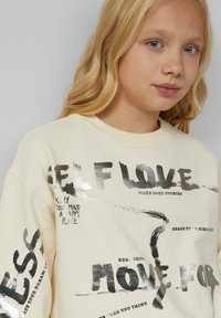 Ragazza bionda con occhi azzurri che indossa un felpa crema con testo motivazionale nero lucido, tra cui "AMORE PER SE STESSI" e "MUOVITI PER".