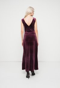 Γυναίκα με μακριά ξανθά μαλλιά, φορώντας ένα αμάνικο βελούδινο φόρεμα σε σκούρο μωβ χρώμα με χαμηλή πλάτη σε V και μαύρες ψηλοτάκουνες γόβες, που στέκεται απέναντι από έναν απλό τοίχο.